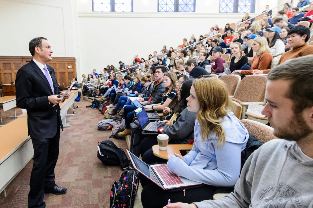 A professor stands before a lecture-style classroom, lecturing to a large group of seated university students.